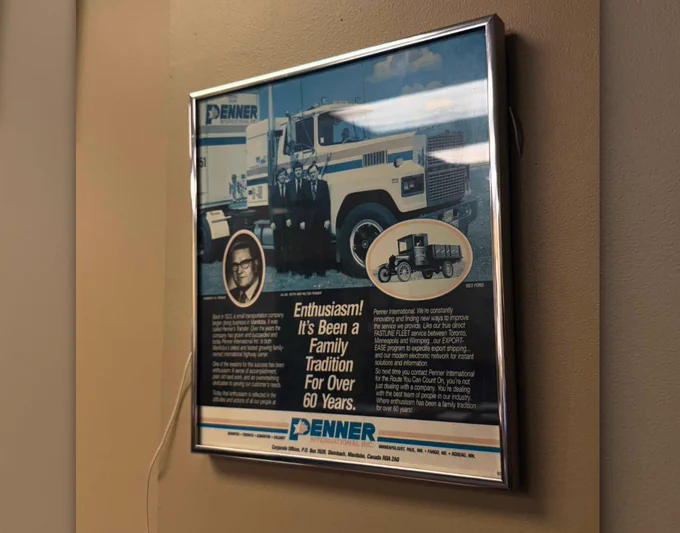 Framed vintage Penner International advertisement hanging on a beige wall. The ad features a large truck with several people standing in front of it, along with smaller images of earlier trucks from the company’s history. The text highlights over 60 years of family tradition and enthusiasm in the transportation industry.