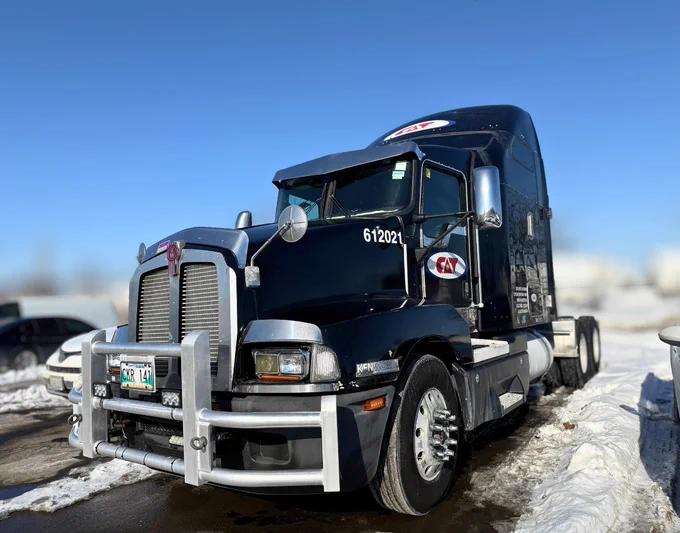 A black C.A.T. Inc. semi‑truck parked outdoors on a sunny winter day, surrounded by snow. The truck has a large chrome grille guard, visible company decals, and a blue sky in the background.