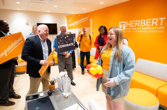 A group of people are gathered in a room at the Herbert College of Agriculture, University of Tennessee, Knoxville. They are celebrating with balloons and signs that say "Congratulations" and "Congrats." The room has orange walls and branding for the college.