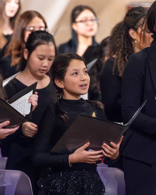 A choir of performers holding black folders, dressed in black attire, sings during a performance. 