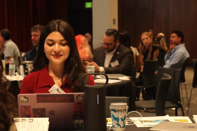 A woman works on her computer. Behind her a mix of men and women sit at round tables  taking notes and talking.

