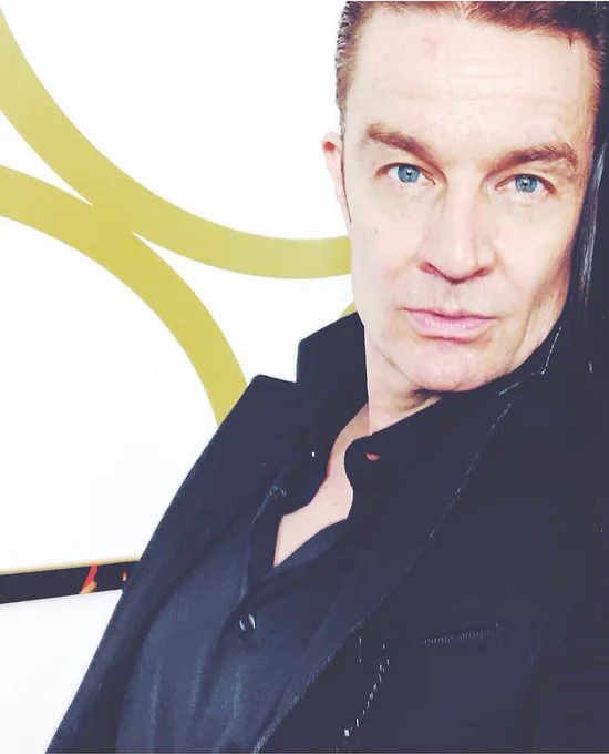 The image features a close-up of a person with short, light brown hair styled back. They have blue eyes and a neutral expression. The person is wearing a black collared shirt and a black jacket. The background is primarily white with an abstract gold design.