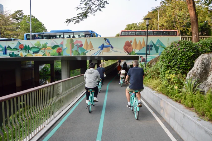 A group of people in casual business attire ride green shared bicycles along dedicated cycling infrastructure through a linear park lined with trees, water, and flowers, in Shenzhen, China.