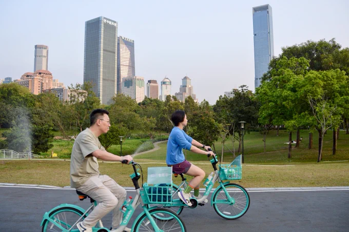 A group of people in casual business attire ride green shared bicycles along dedicated cycling infrastructure through a linear park lined with trees, water, and flowers, in Shenzhen, China.