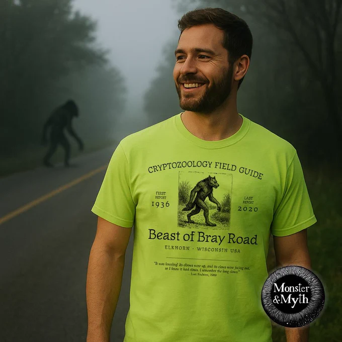 A man wearing a bright green “Beast of Bray Road” cryptozoology field guide T-shirt stands smiling on a foggy rural road, while a tall shadowy cryptid figure resembling a dogman or werewolf looms indistinctly in the mist behind him among the trees.
