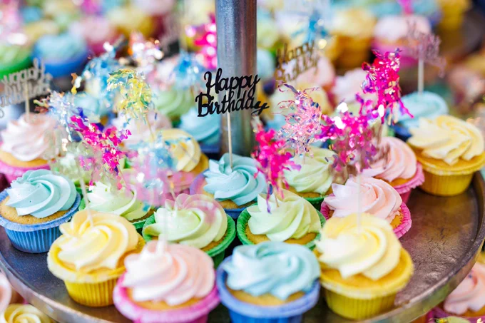 A display of colorful cupcakes with swirled frosting in pastel shades, each topped with decorative picks. Some picks have the words "Happy Birthday" while others feature shiny, metallic decorations.