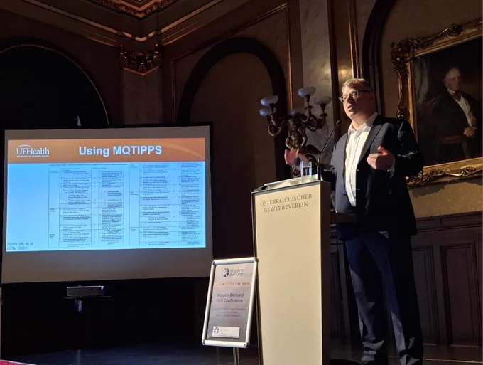 Philip Efron giving a talk at a podium. In the background, a presentation slide is headed with "Using MQTIPPS."
