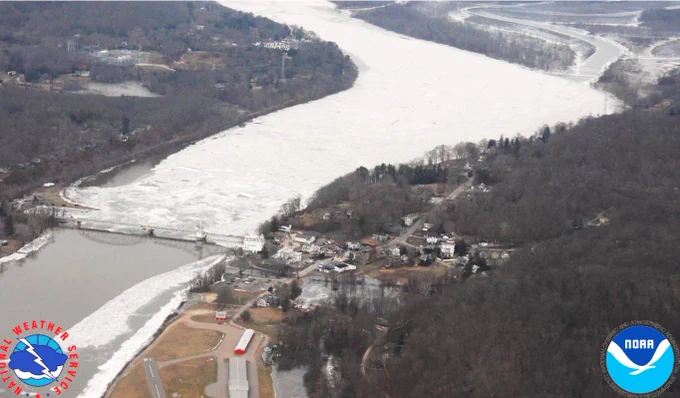 Aerial view of a frozen river choked with ice floes, passing under a bridge near a small town and forested hills. NOAA and NWS logos in corners.