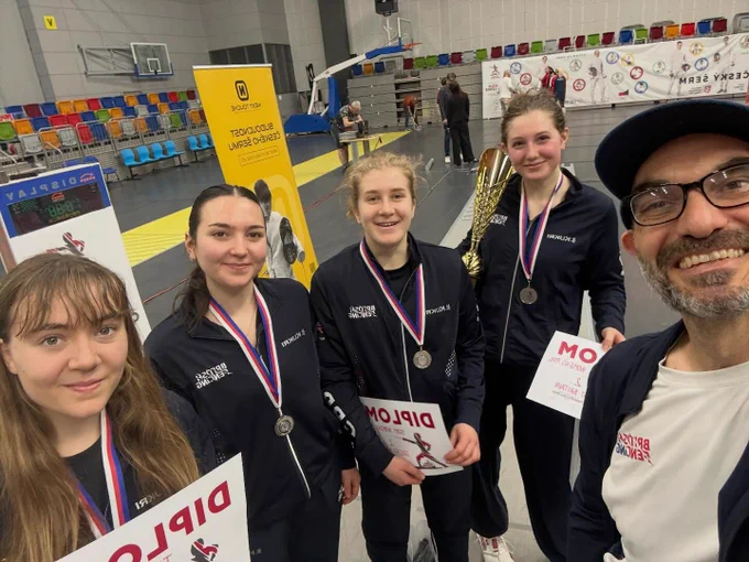 A selfie of four GBR foil fencers smiling while holding up certificates and medals, alongside their coach. One is holding a trophy.