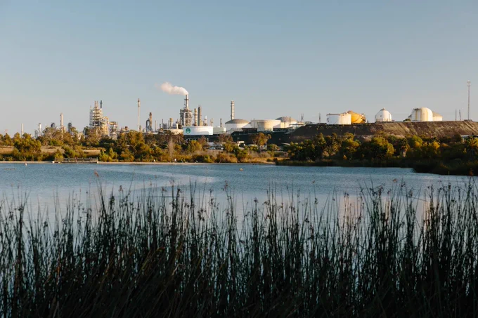 Industrial refinery complex with storage tanks and smokestacks emitting steam, viewed across a calm body of water with reeds in the foreground. Courtesy of CalMatters