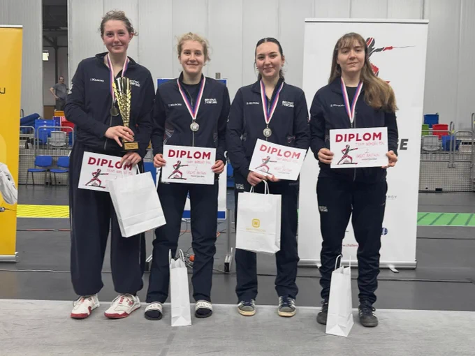 Four GBR fencers stand smiling to the camera, wearing medals and holding certificates. They are holding a trophy.