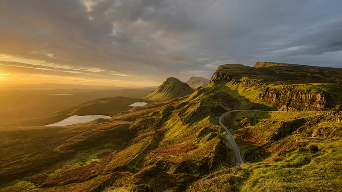Sunset casting golden light over rolling green hills and winding road in a rugged mountainous landscape.