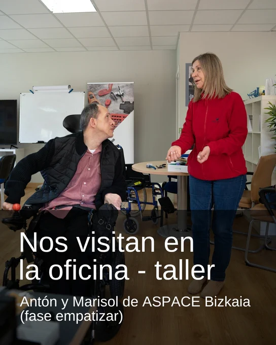 Persona en silla de ruedas conversando con otra persona de pie en un espacio de trabajo. Texto sobreimpreso: “Nos visitan en la oficina - taller. Antón y Marisol de ASPACE Bizkaia (fase empatizar)”.
