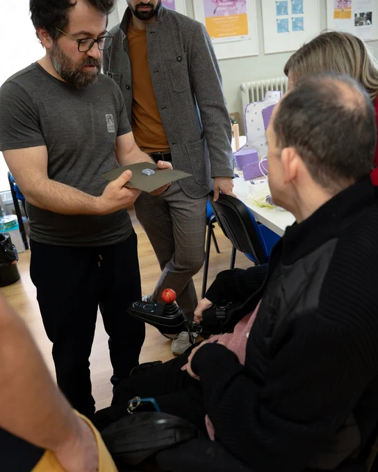 Persona mostrando una pieza impresa en 3D sobre una base a otra persona en silla de ruedas, con varias personas observando.