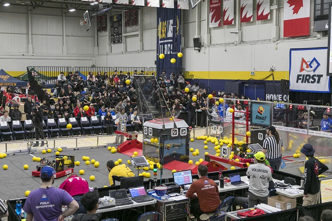 Robots in an arena are shooting balls into a container as people watch.
