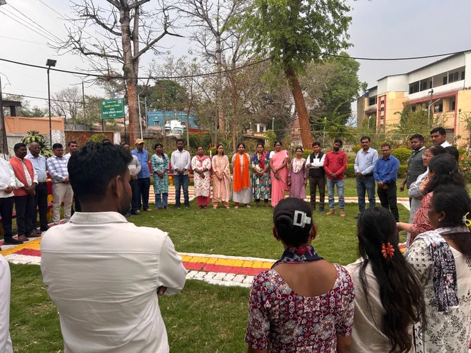 A group of people stand in a wide circle outdoors on a grassy area, appearing to participate in a group activity.

