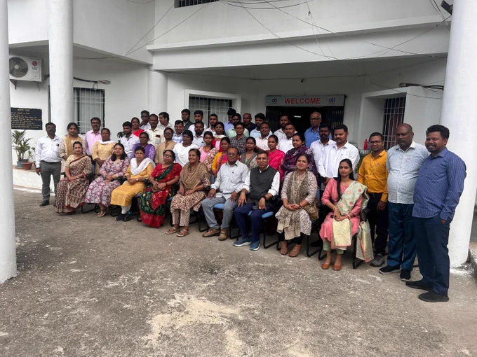 A large group of adults pose for a group photo outside a white building, seated and standing in rows near an entrance with a “WELCOME” sign.
