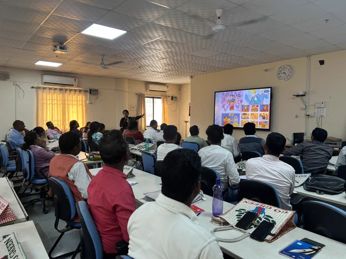 A classroom training session with participants seated at desks, watching a presenter speak beside a screen displaying illustrated slides.
