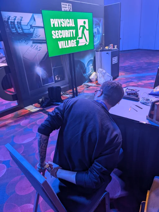 A man handcuffed, sitting on a chair, with the Physical Security Village logo on a tv in the background