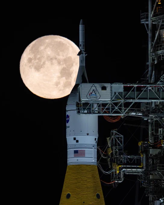 A full Moon appears large and bright in the night sky behind a NASA rocket on its launch pad. The rocket\u2019s white upper stage, orange lower section, and surrounding launch tower structure are illuminated against the dark background.