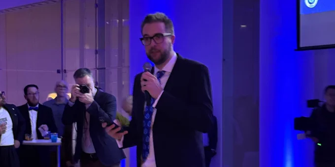 Man in a suit and sneakers speaking into a microphone at a formal event with blue lighting and an audience in the background.