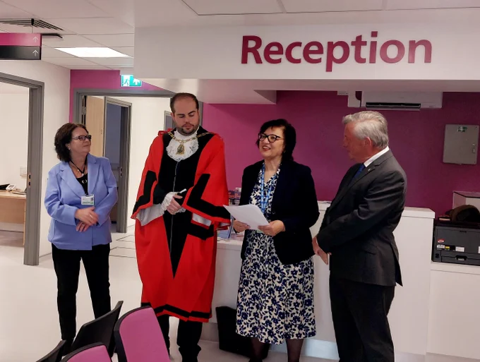 A group of people including NHS collegaues, Bromley Council colleagues and the Mayor of Bromley standing inside the Bromley Health and Wellbeing Centre. Dr Angela Bhan is reading a speech. The group stands in a modern reception area with signage for Bromley Health and Wellbeing Centre in the background.