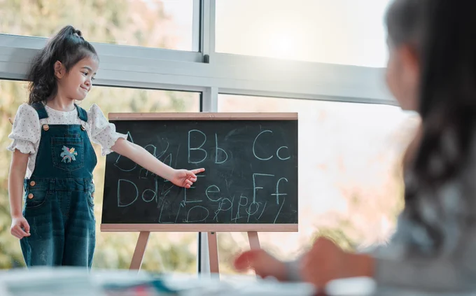 Jeune enfant debout devant un petit tableau noir, pointant des lettres de l’alphabet écrites à la craie, tandis qu’un adulte est assis en arrière-plan, légèrement flou, dans un environnement lumineux de type salle de classe ou espace d’apprentissage.