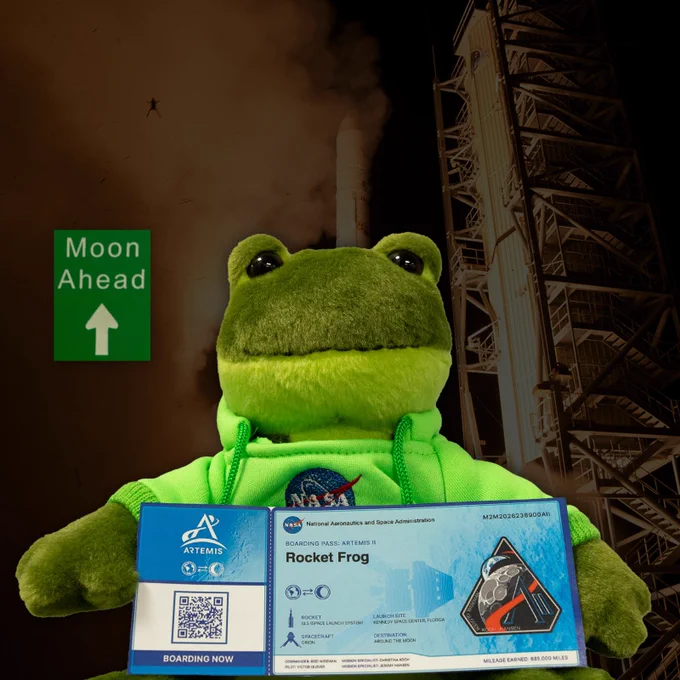 A plush green frog holding a blue Artemis II boarding pass between its arms. The boarding pass has the name Rocket Frog on it. To the left of the frog is a white and green road sign with the words \"Moon Ahead\" and a up arrow on it. The background is a faded into black image of the LADEE launch with a frog silhouette. 