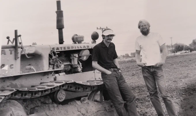 Two individuals are standing beside a Caterpillar bulldozer in an open, muddy field. The person on the left is wearing a cap and glasses, and the one on the right has crossed arms. There is a telephone pole and trees in the background. The image is in black and white.