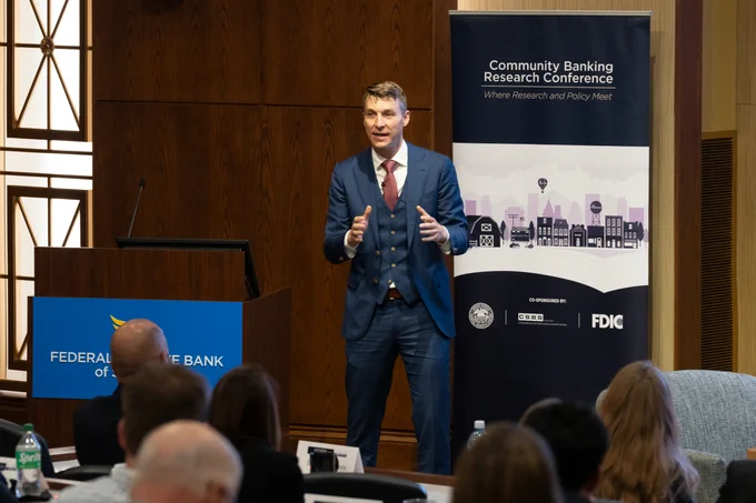 A person speaks at a podium, addressing an audience. The banner behind reads "Community Banking Research Conference."