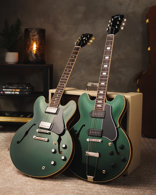 Two Gibson British Racing Green electric guitars with semi-hollow bodies are leaning against a beige amplifier. The setting includes a shelf with books and a small plant, alongside a soft-lit environment.