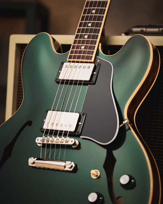 A close-up of a British Racing Green semi-hollow electric guitar, resting against an amplifier. The guitar features dual humbucker pickups, a stylish black pickguard, and elegant cream binding along the edges.