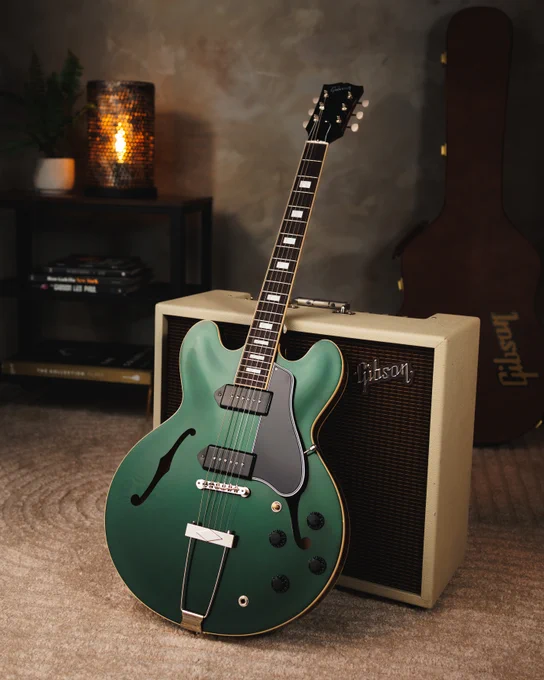 A British Racing Green semi-hollow electric guitar is leaning against a cream-colored Gibson amplifier. In the background, there's a black shelf with books and a lit candle. A guitar case is also visible.