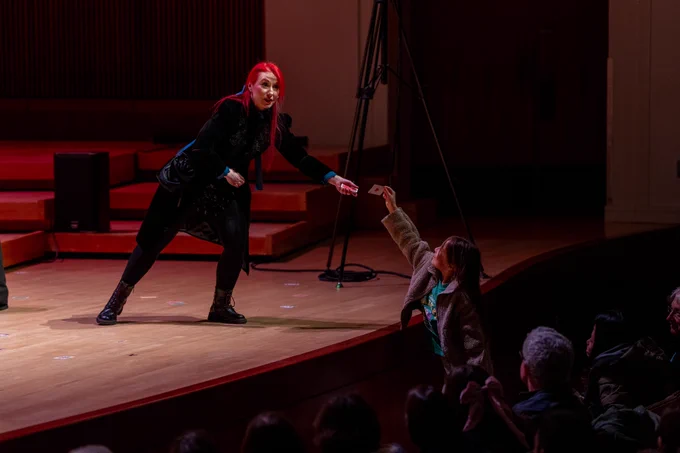 A woman with red hair standing on a stage getting a card from an audience member for a card trick