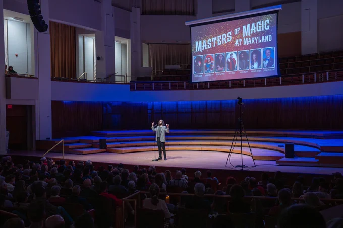 A man standing on a stage behind a microphone with a screen behind him that reads "Masters of Magic at Maryland"
