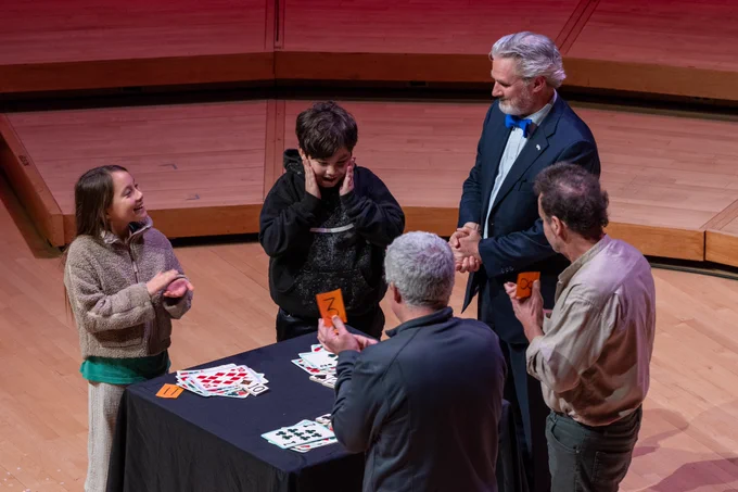 A performer stands on stage at a table with different cards and four audience members