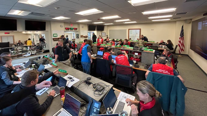 A group of about 30 people in a room sitting around three tables working on computers and talking.