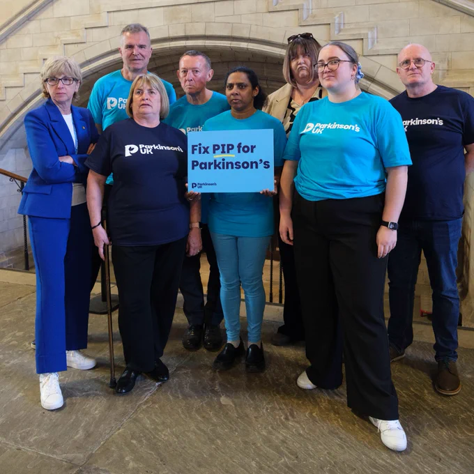 A group of adults are standing together wearing Parkinson's UK t-shirts in various shades of blue. Our CEO Caroline has joined them in a blue suit. Everyone looks serious as they hold a placard that reads 'Fix PIP for Parkinson's'