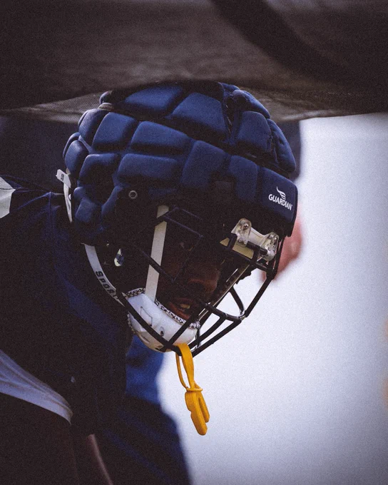 Close-up profile of a football player wearing a navy blue helmet with a padded Guardian Cap and a yellow mouthguard hanging from the face mask.