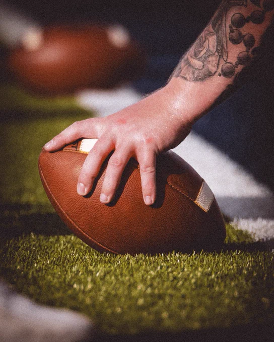 Extreme close-up of a tattooed hand gripping a football on the turf, with another football blurred in the background.