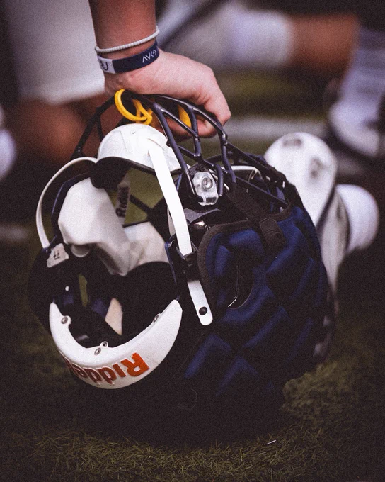 A close-up shot of a hand holding a white Riddell football helmet by the face mask, featuring a navy blue Guardian Cap and a yellow mouthguard.