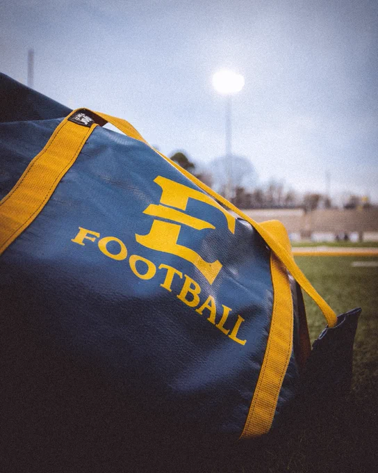 A low-angle shot of a blue and gold equipment bag on the grass with the E FOOTBALL logo prominently displayed under bright stadium lights.