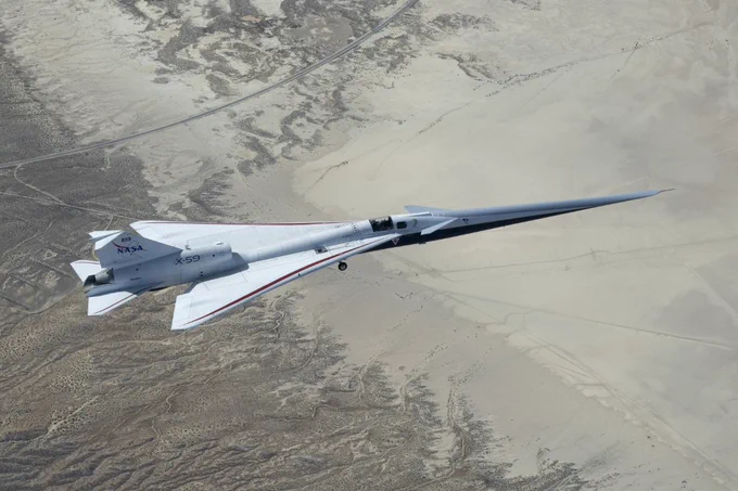 NASA\u2019s white, blue, and red X-59 research aircraft flies over the Mojave Desert near NASA\u2019s Armstrong Flight Research Center in Edwards, California, during its third flight. The aircraft is centered in the frame, viewed from above with the canopy, both wings, NASA and X-59 logos visible, and the landing gear down with one wheel visible. Below, rugged terrain transitions into the smooth texture of a dry lakebed as the aircraft, nearly 100 feet long, flies toward the right side of the frame.