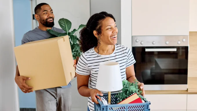 Two people are smiling and carrying items in a kitchen. One person holds a cardboard box and a potted plant, while the other carries a blue basket containing a lamp and a small plant. A modern oven is visible in the background.