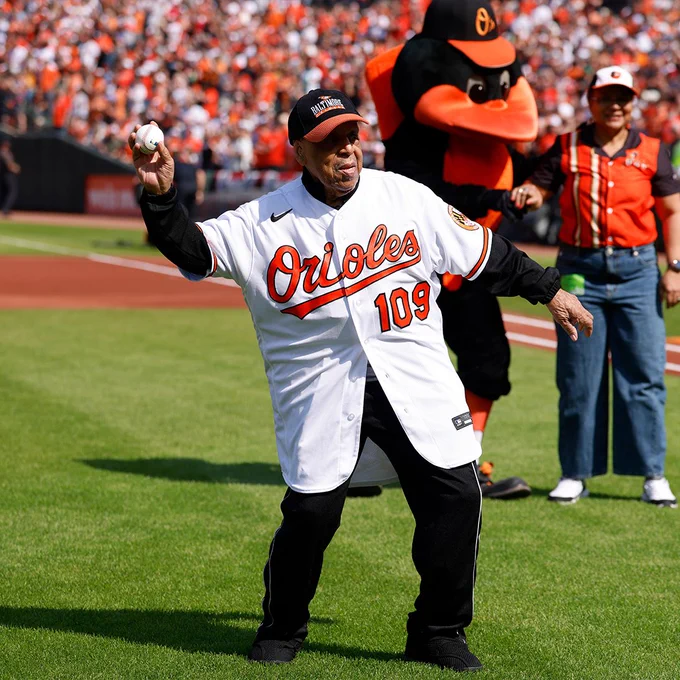 109-year-old Arthur Green throws out the ceremonial first pitch at Baltimore Orioles Opening Day. He wears a white Orioles jersey with the number 109 on the front and a black and orange Orioles cap. The Oriole Bird is seen in the background.