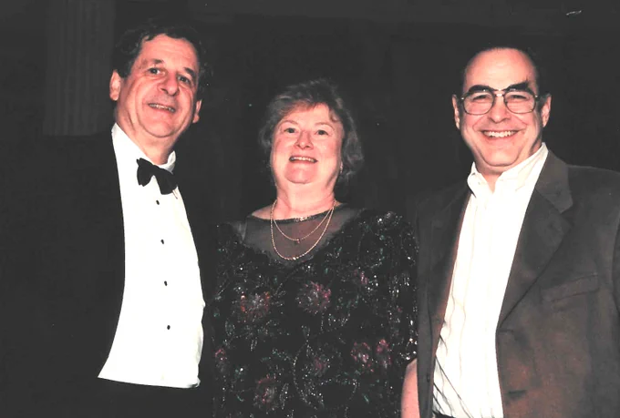 Three people in formalwear. The woman in the middle is wearing a dress with roses on it.