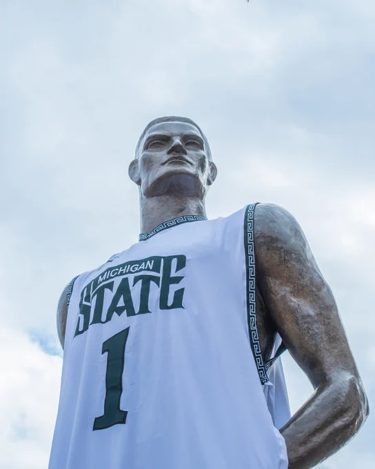 Low-angle shot of the Spartan statue wearing a white Michigan State jersey with the number 1. The statue faces forward with a cloudy sky behind it.