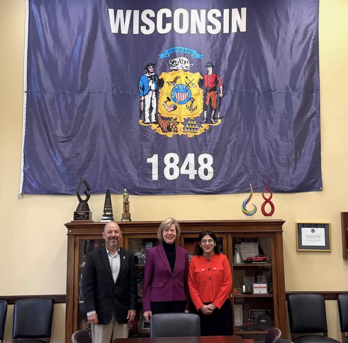 Three individuals standing in front of a large Wisconsin state flag in an interior room
