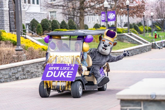 Duke Dog rides a golf cart decorated with streamers and a sign that reads “Give Like a Duke”.