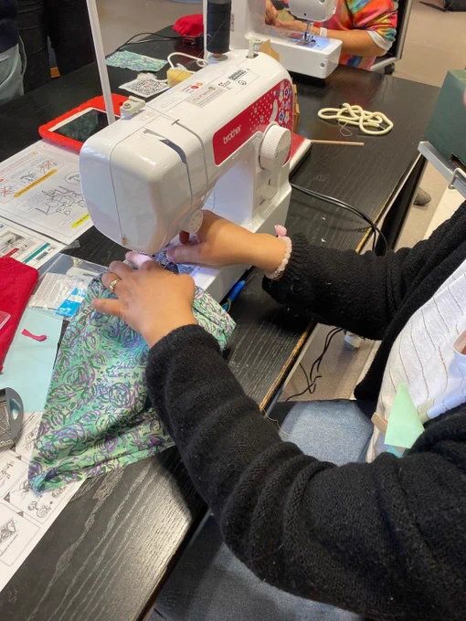 Personne en train d'utiliser une machine à coudre posée sur une table, cousant un tissu à motifs verts et bleus. Autour, on voit des feuilles de papier et des croquis, du matériel de couture et une autre machine à coudre en arrière-plan. 